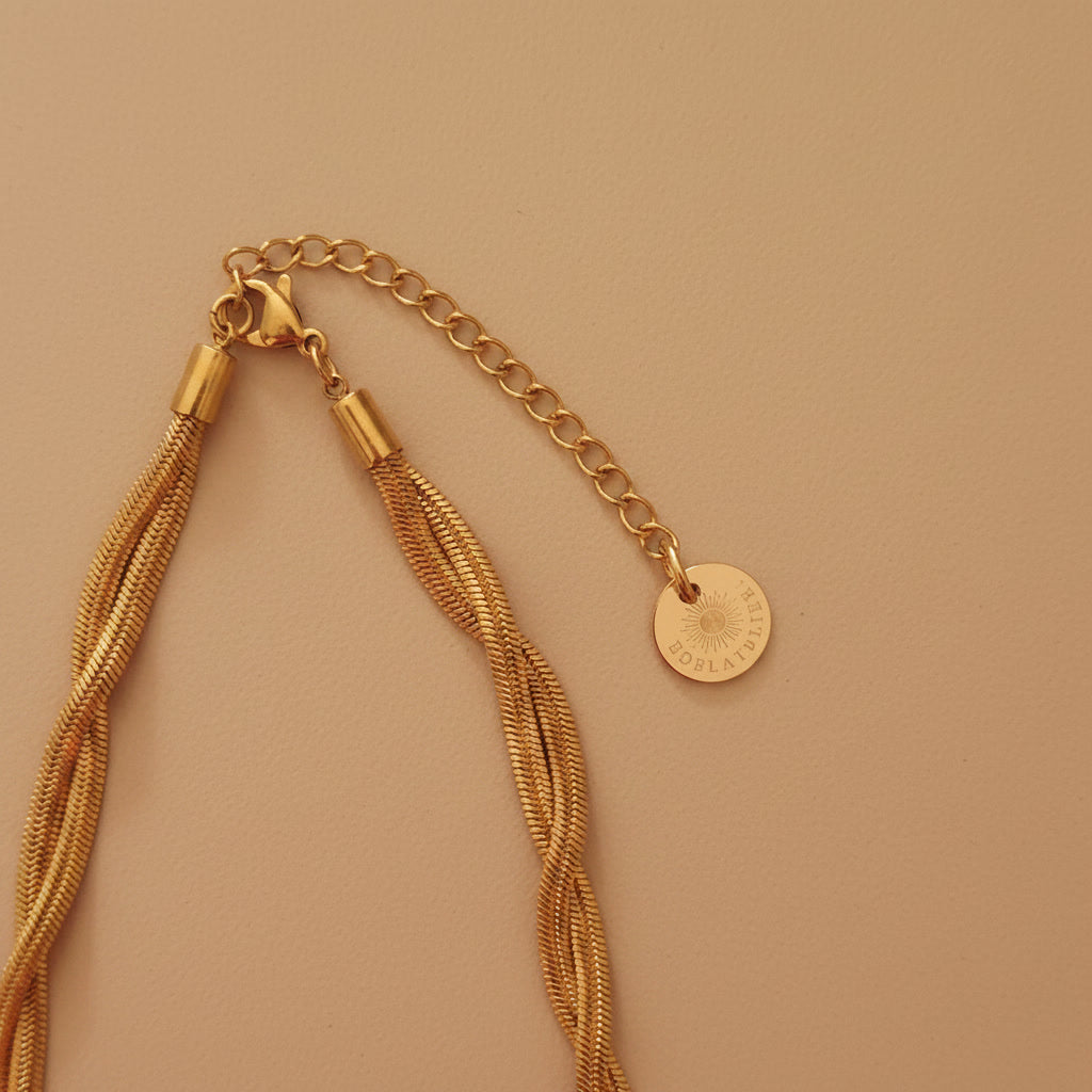 Detailaufnahme der goldfarbenen Intreccia Halskette mit Karabiner, Verlängerungskette und EDELATELIER.-Logoplättchen.
