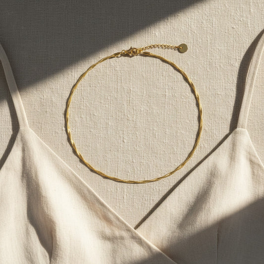 Goldfarbene Intreccia Halskette mit gedrehter Struktur auf hellem Leinenkleid im Sonnenlicht.