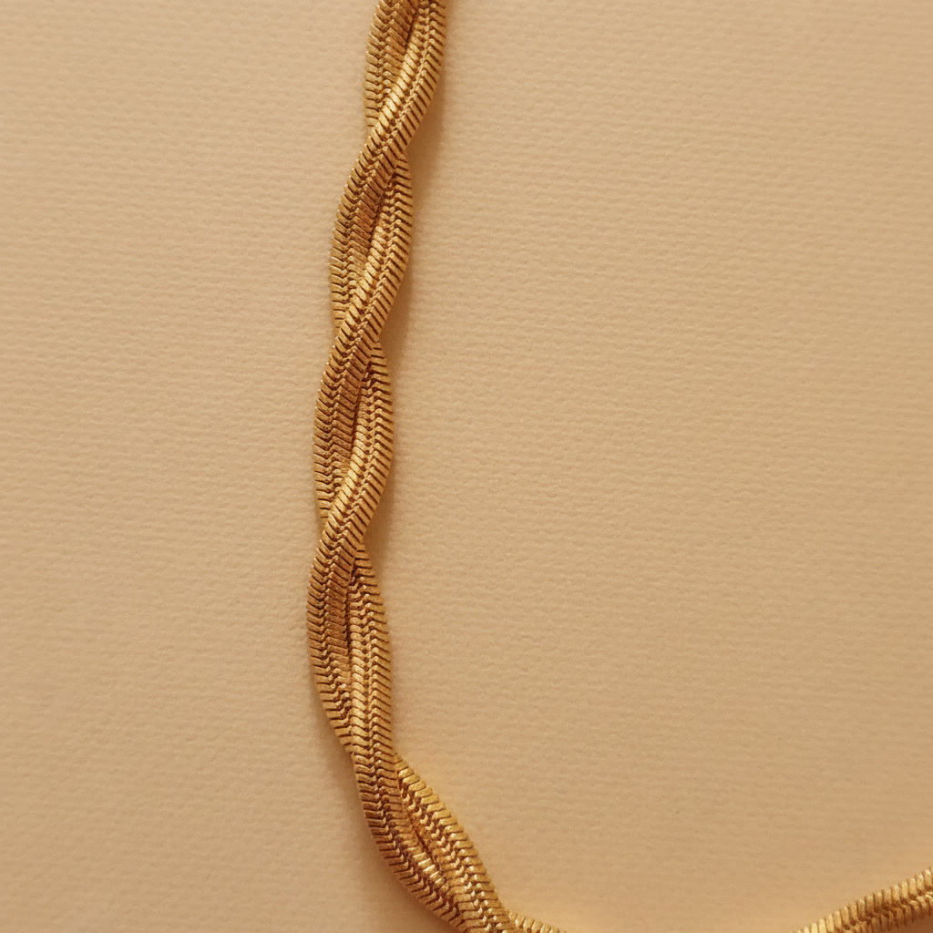 Goldfarbene Intreccia Halskette mit gedrehter, mehrsträngiger Struktur auf warmem beigem Hintergrund.