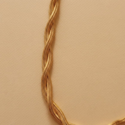 Goldfarbene Intreccia Halskette mit gedrehter, mehrsträngiger Struktur auf warmem beigem Hintergrund.
