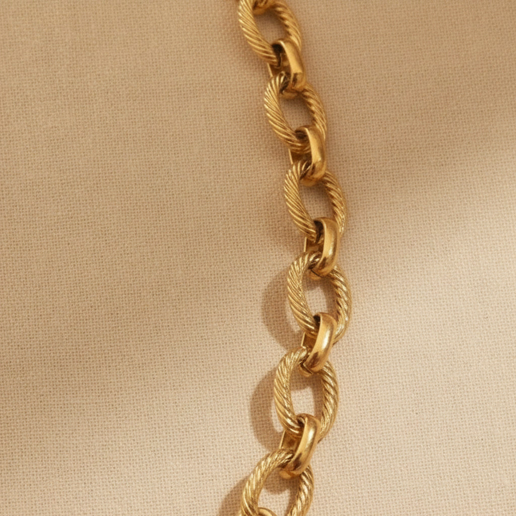 Goldfarbene Maglia Halskette mit ovalen strukturierten Gliedern, rundem Frontverschluss und Logoplättchen auf warmem beigem Leinenhintergrund.