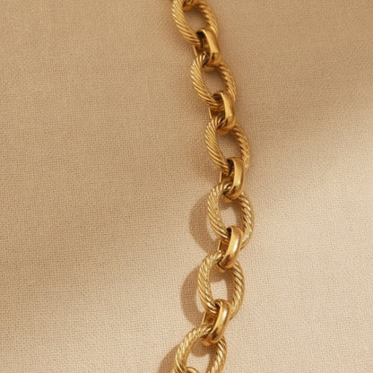 Goldfarbene Maglia Halskette mit ovalen strukturierten Gliedern, rundem Frontverschluss und Logoplättchen auf warmem beigem Leinenhintergrund.