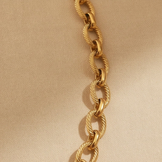 Goldfarbene Maglia Halskette mit ovalen strukturierten Gliedern, rundem Frontverschluss und Logoplättchen auf warmem beigem Leinenhintergrund.