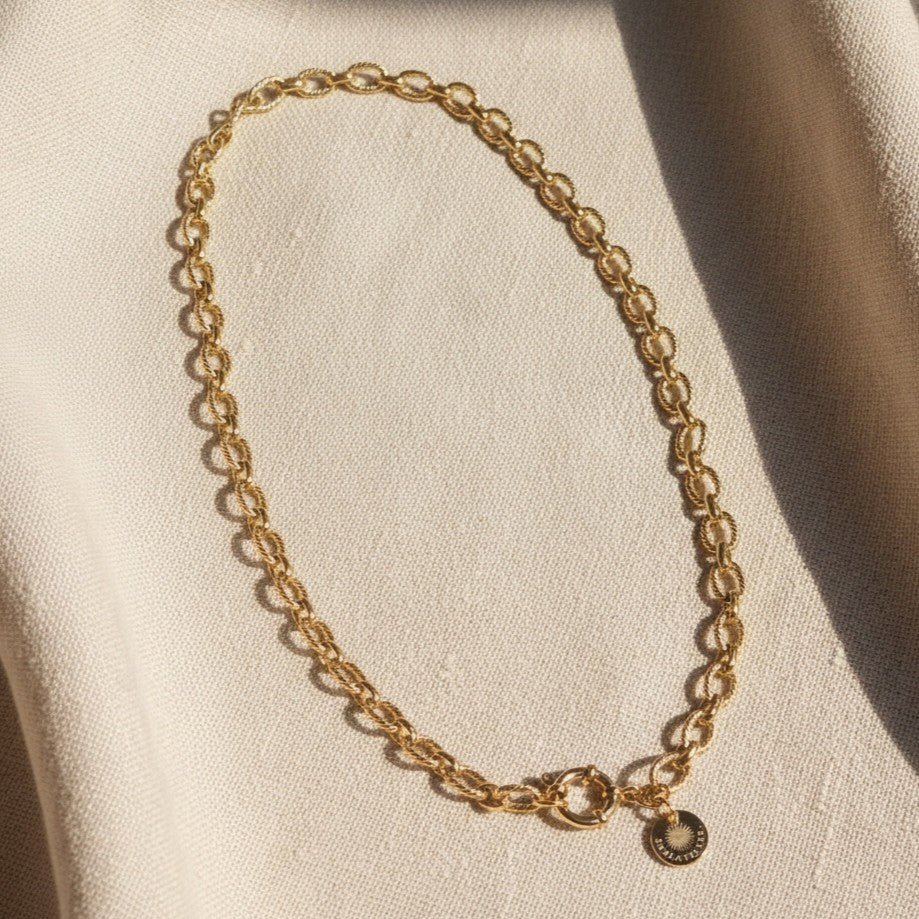 Goldfarbene Maglia Halskette mit ovalen Gliedern, rundem Verschluss und EDELATELIER.-Logoplättchen auf warmem beigem Leinenhintergrund.
