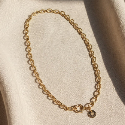 Goldfarbene Maglia Halskette mit ovalen Gliedern, rundem Verschluss und EDELATELIER.-Logoplättchen auf warmem beigem Leinenhintergrund.