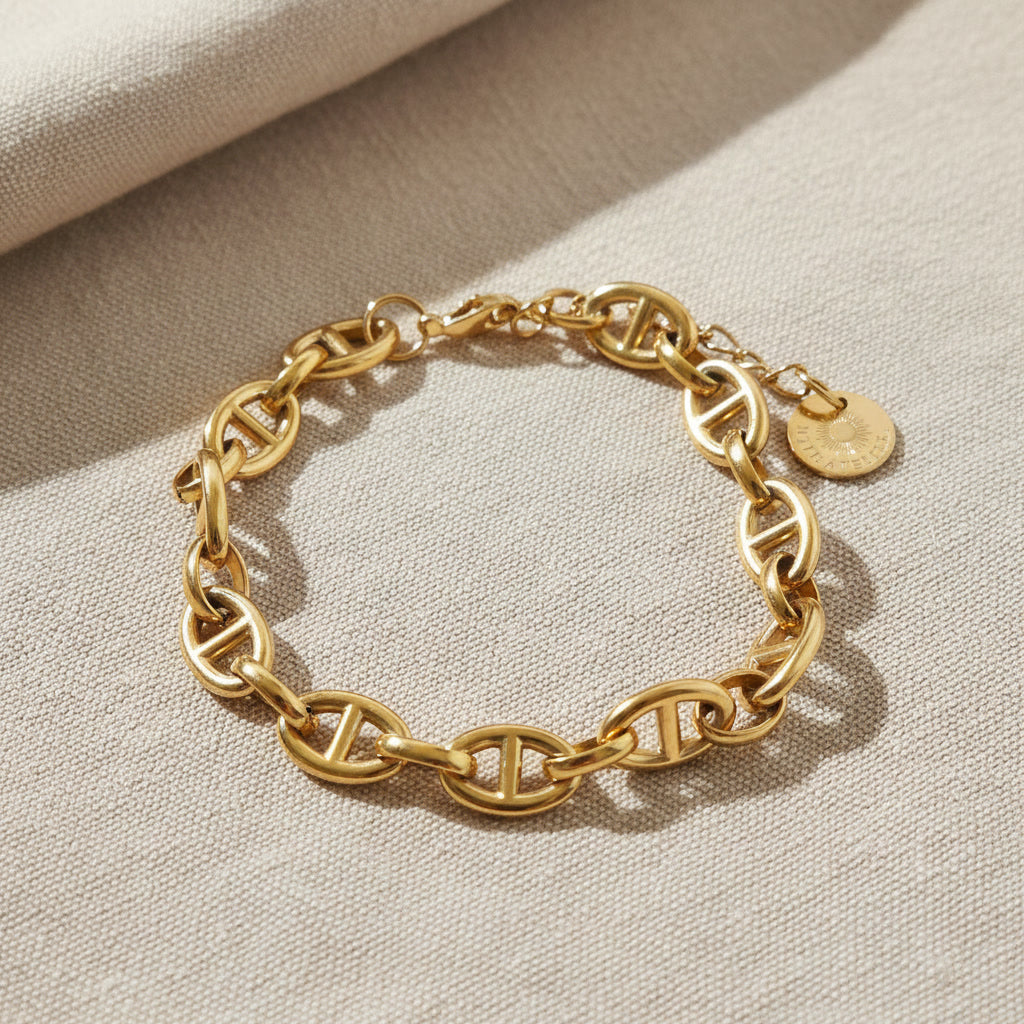 Marea Gliederarmband aus goldfarbenem Edelstahl auf beigem Leinen, markante Kettenglieder und Logo-Anhänger sichtbar.
