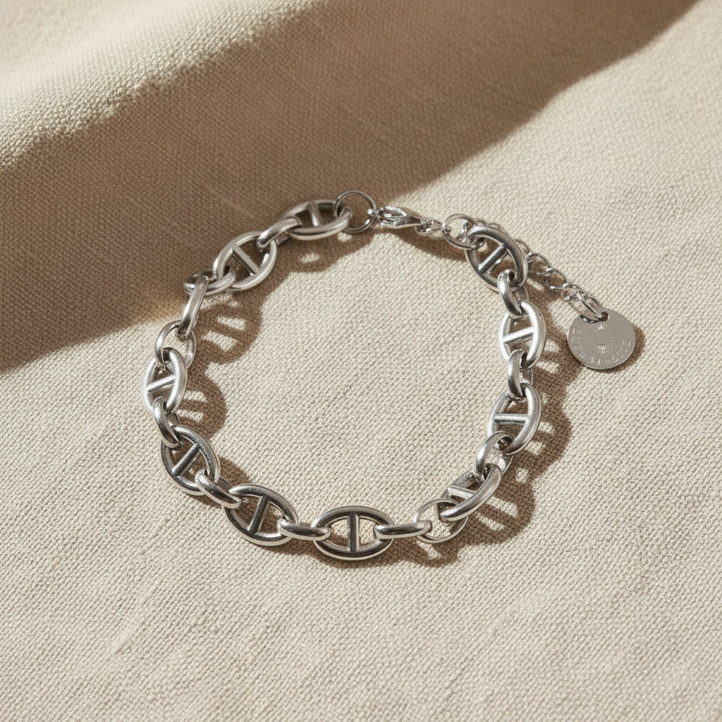 Marea Gliederarmband aus silberfarbenem Edelstahl auf beigem Leinen, markante Kettenglieder und Logo-Anhänger sichtbar.