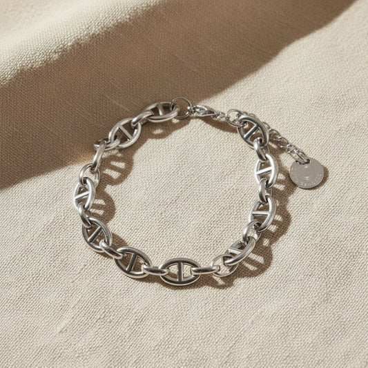 Marea Gliederarmband aus silberfarbenem Edelstahl auf beigem Leinen, markante Kettenglieder und Logo-Anhänger sichtbar.