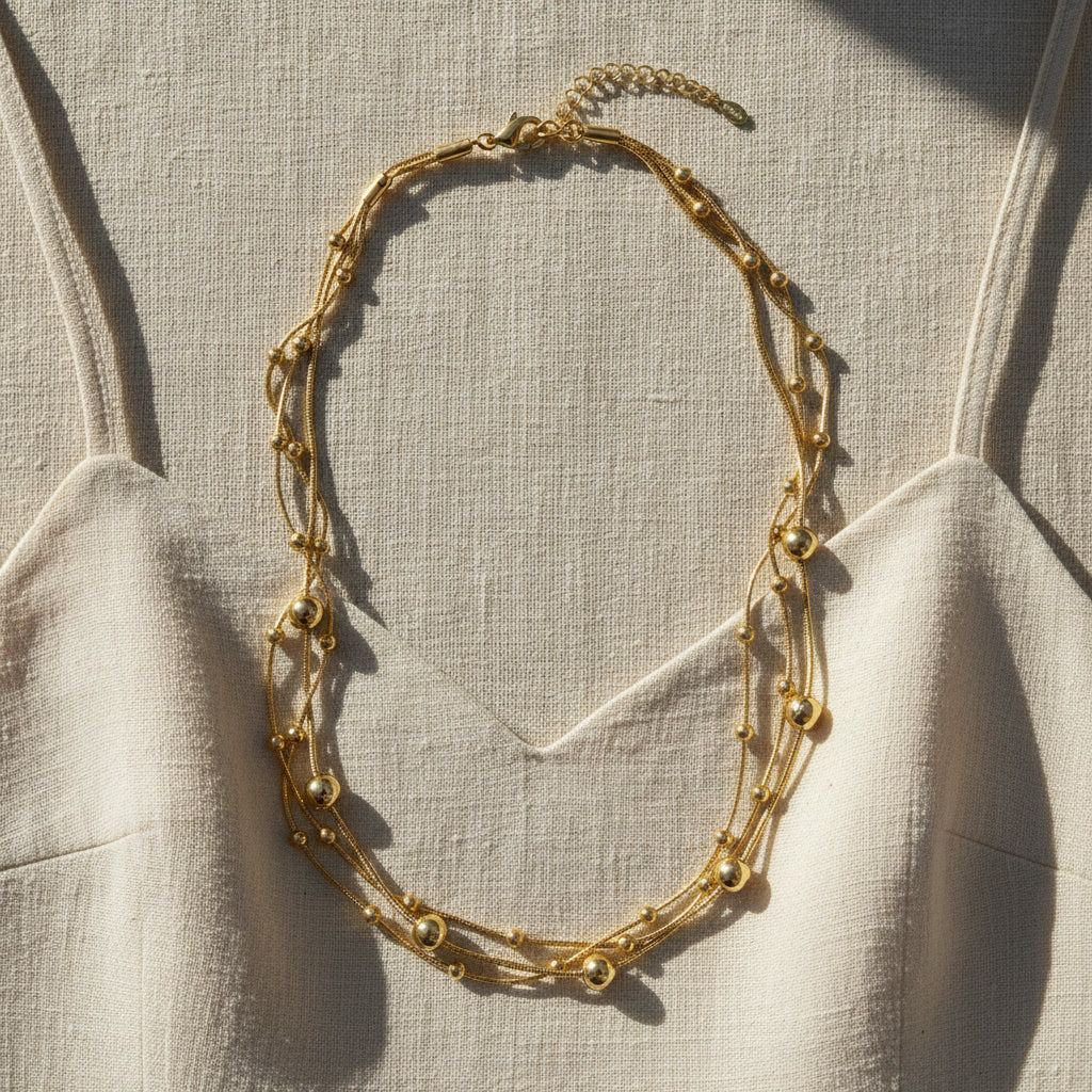 Mehrreihige goldfarbene Halskette mit Kugeldetails auf hellem Leinen-Top im warmen Sonnenlicht.