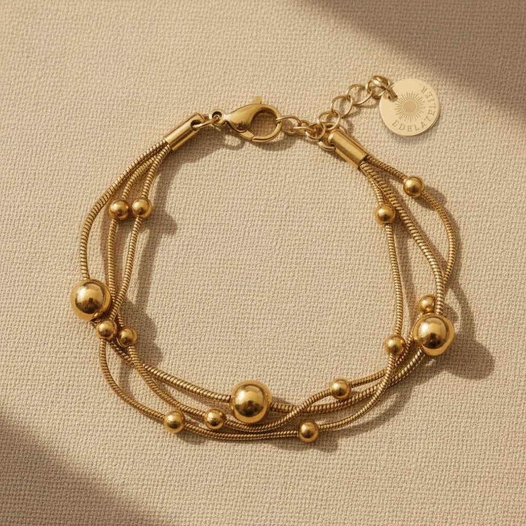 Goldfarbenes Piccola Perla Armband mit feinen Edelstahlsträngen und glänzenden Kugeldetails auf warmem beigem Hintergrund.