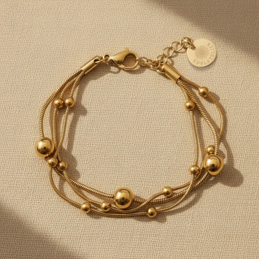 Goldfarbenes Piccola Perla Armband mit feinen Edelstahlsträngen und glänzenden Kugeldetails auf warmem beigem Hintergrund.