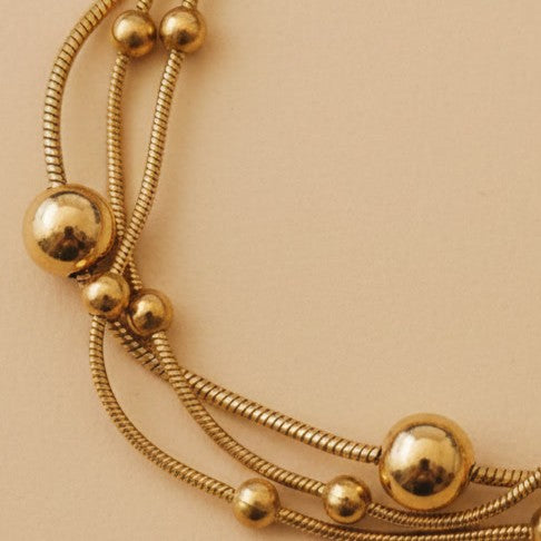 Detailaufnahme des goldfarbenen Piccola Perla Armbands mit glänzenden Kugeln und feiner Edelstahlstruktur auf warmem beigem Hintergrund.