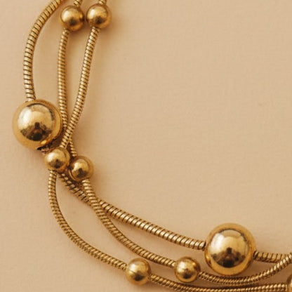 Detailaufnahme des goldfarbenen Piccola Perla Armbands mit glänzenden Kugeln und feiner Edelstahlstruktur auf warmem beigem Hintergrund.
