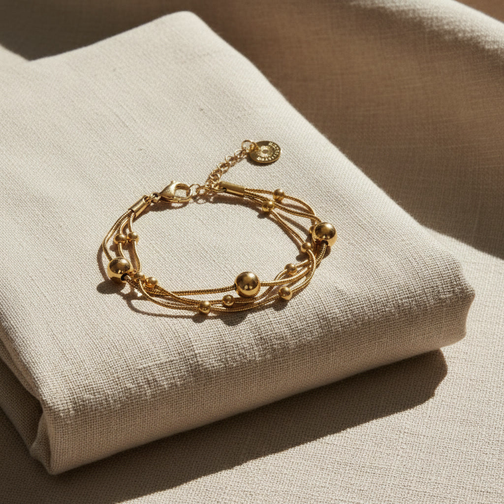 Goldfarbenes Piccola Perla Armband mit feinen Kugeldetails auf gefaltetem beigem Leinenstoff im warmen Sonnenlicht.