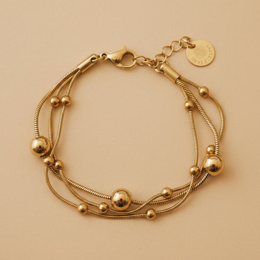 Goldfarbenes Piccola Perla Armband mit feinen Edelstahlsträngen und glänzenden Kugeldetails auf warmem beigem Hintergrund.