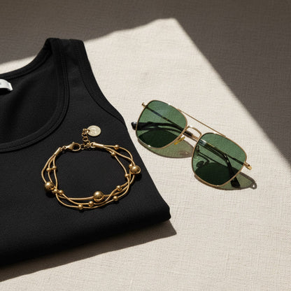 Goldfarbenes Piccola Perla Armband auf schwarzem Top neben grüner Sonnenbrille im warmen Sonnenlicht arrangiert.