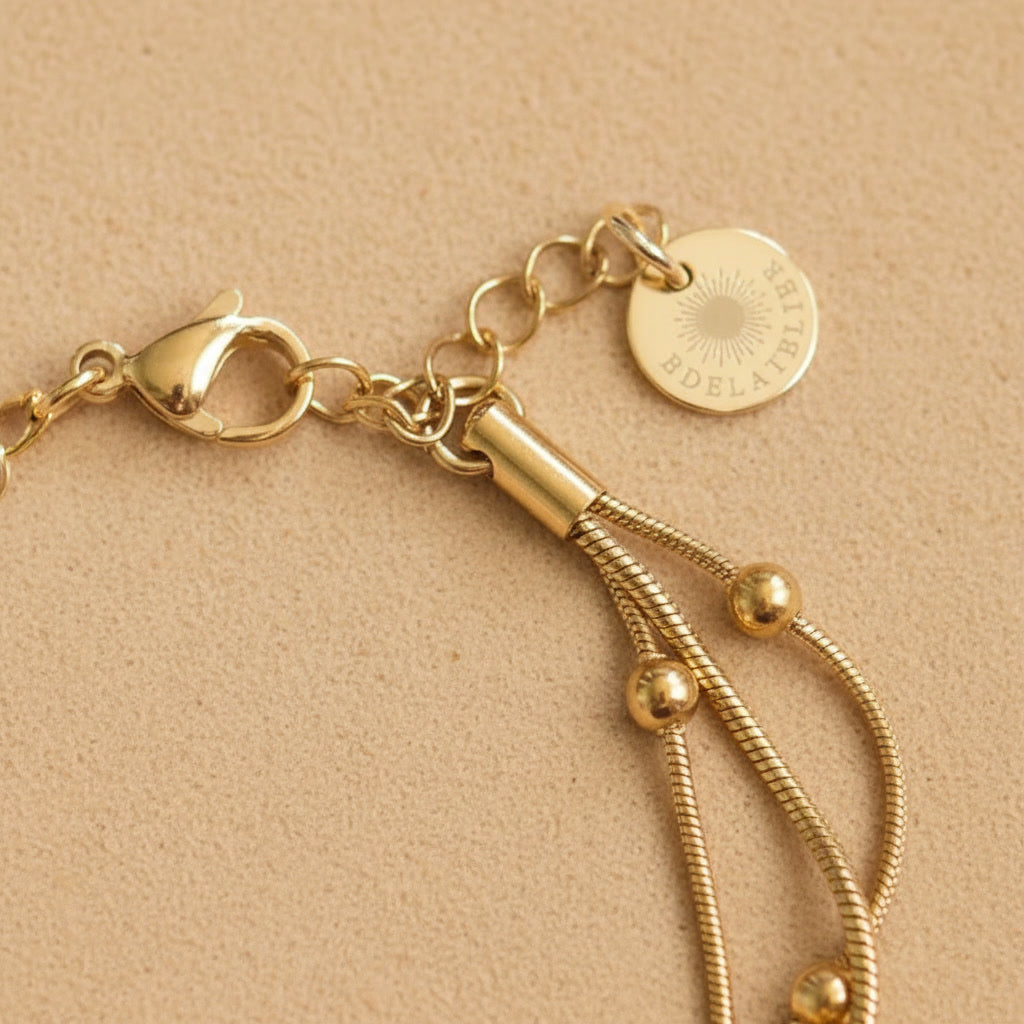 Detailaufnahme des goldfarbenen Piccola Perla Armbands mit Verlängerungskette und EDELATELIER.-Logoplättchen auf warmem beigem Hintergrund.