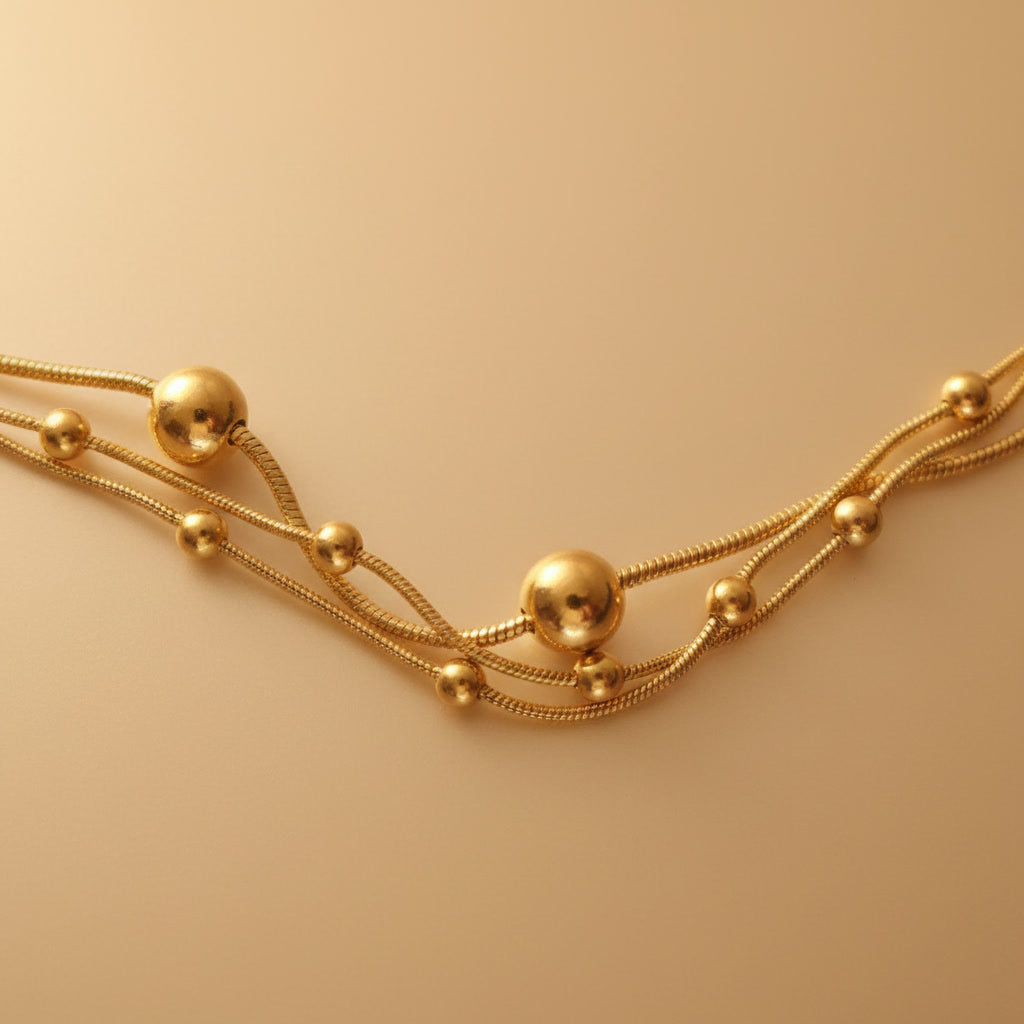 Detailaufnahme der goldfarbenen Piccola Perla Halskette mit feinen Kettensträngen und kleinen Kugeln auf warmem beigem Hintergrund.
