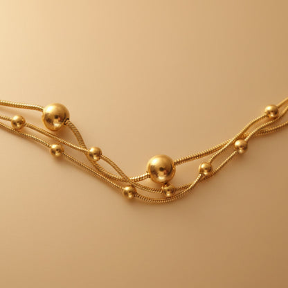 Detailaufnahme der goldfarbenen Piccola Perla Halskette mit feinen Kettensträngen und kleinen Kugeln auf warmem beigem Hintergrund.