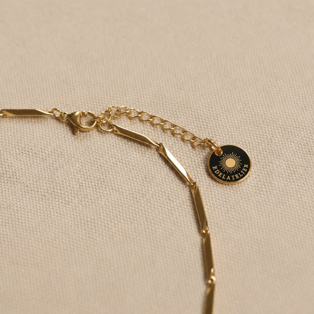 Detailaufnahme der goldfarbenen Saetta Halskette aus Edelstahl mit Karabinerverschluss, Verlängerungskette und EDELATELIER-Logoplättchen auf hellem Leinenstoff.