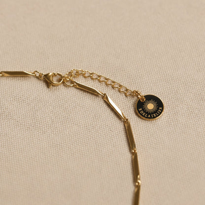 Detailaufnahme der goldfarbenen Saetta Halskette aus Edelstahl mit Karabinerverschluss, Verlängerungskette und EDELATELIER-Logoplättchen auf hellem Leinenstoff.