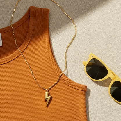goldfarbene Saetta Blitz-Halskette mit Anhänger auf eiem orangenem Tanktop, nebem einer gelben Sonnenbrille.