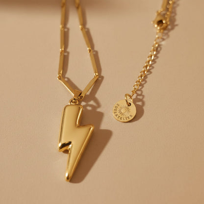 Detailaufnahme der goldfarbenen Saetta Halskette mit glänzendem Blitzanhänger, feiner Kette und warmem beigem Hintergrund.