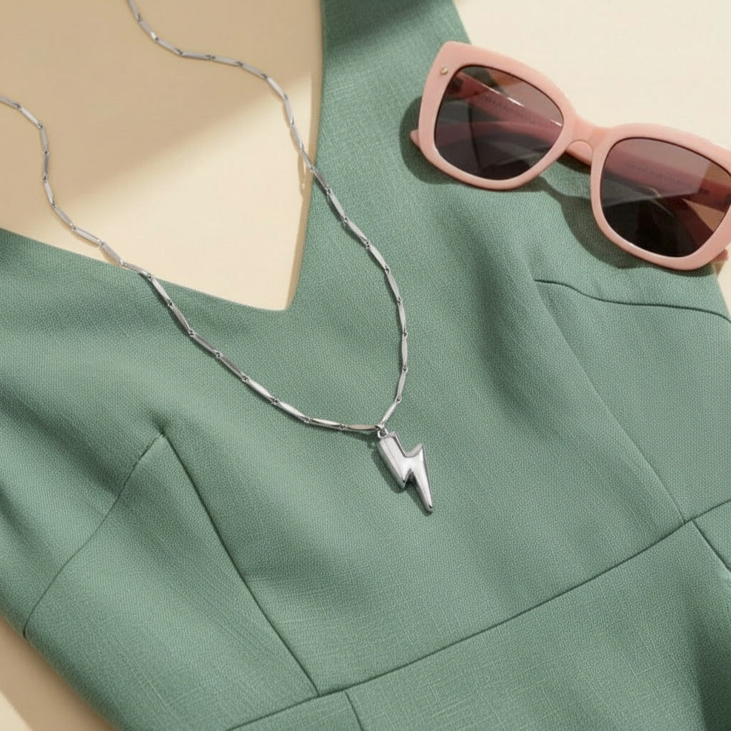 Silberfarbene Saetta Halskette mit Blitzanhänger auf mintgrünem Kleid, kombiniert mit rosafarbener Sonnenbrille.