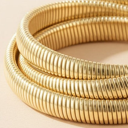 Detailaufnahme des goldfarbenen Spirale d’Oro Armbands mit gerippter Edelstahl-Struktur auf warmem beigem Hintergrund.