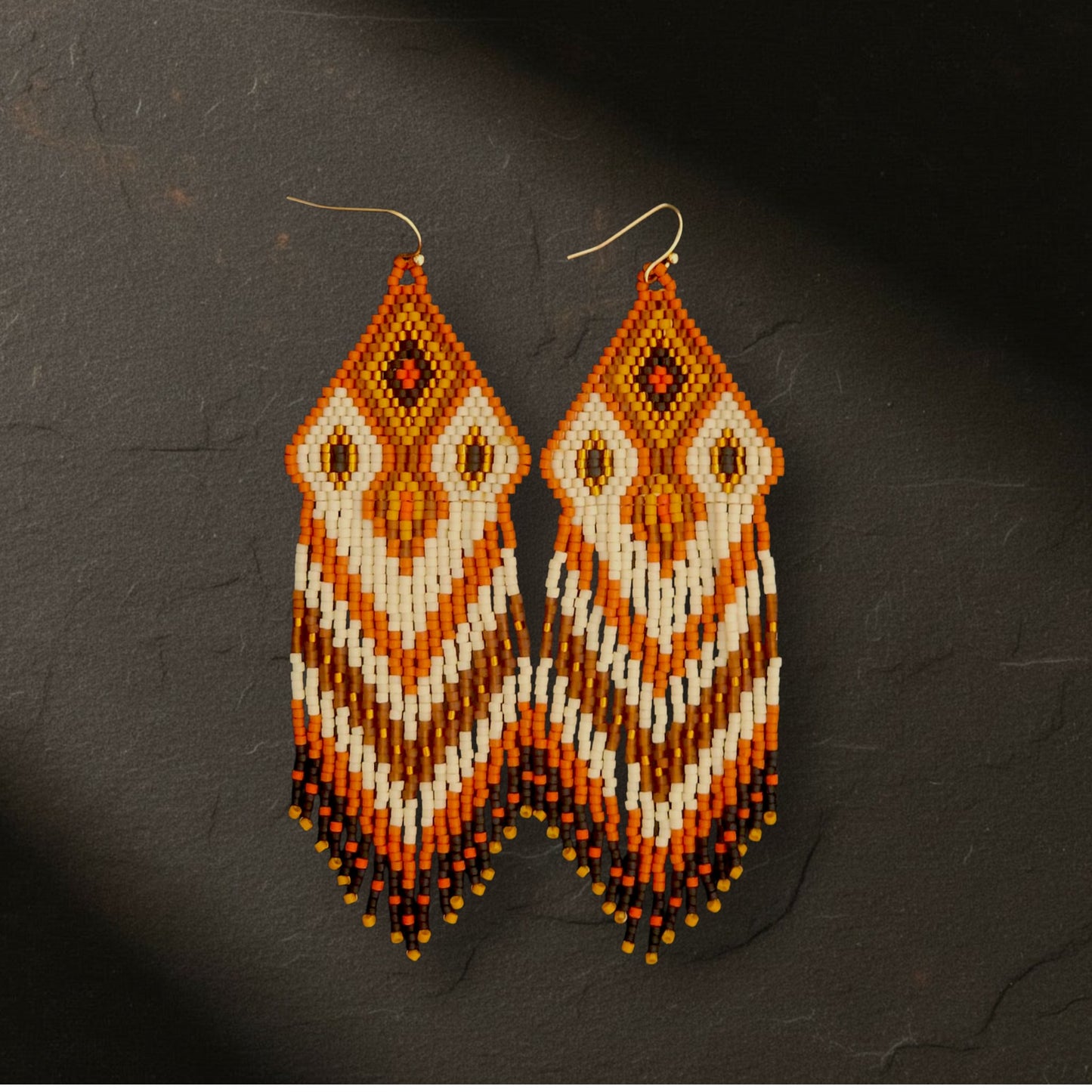 Ein Paar handgefertigte Statement-Ohrringe Modell Fiamma di Terra von EDELATELIER, präsentiert auf dunklem Naturschiefer. Die 10 cm langen Ohrringe zeigen ein warmes, erdnahes Muster in kräftigem Rostrot und Terrakotta, kombiniert mit cremeweißen und braunen Akzenten sowie feinen Golddetails. Die filigranen Perlenfransen und das präzise Webmuster kommen auf dem rustikalen Steinuntergrund besonders kontrastreich zur Geltung.
