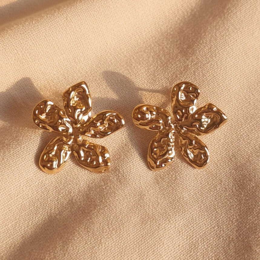 Fiorella Edelstahlohrringe in Blumenform aus Gold auf einem warmbeigen Leinenuntergrund. 