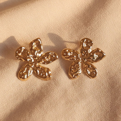 Fiorella Edelstahlohrringe in Blumenform aus Gold auf einem warmbeigen Leinenuntergrund. 