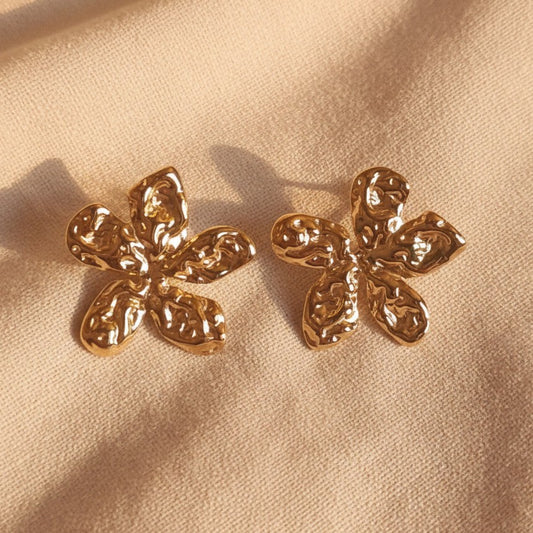 Fiorella Edelstahlohrringe in Blumenform aus Gold auf einem warmbeigen Leinenuntergrund. 