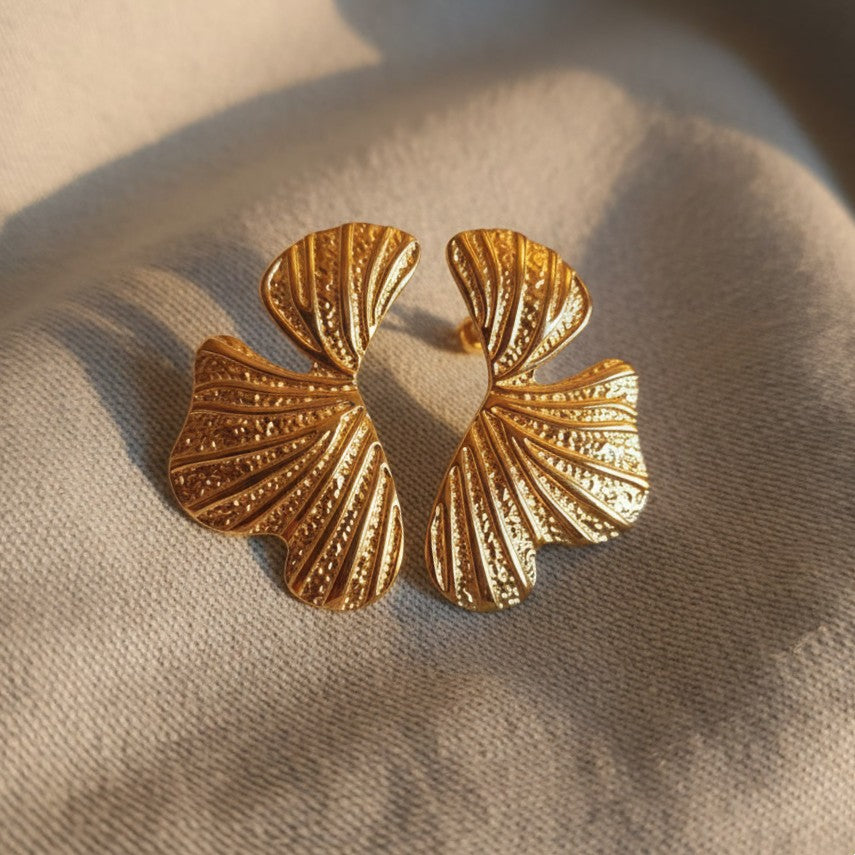 Goldfarbene Foglia Ohrringe aus Edelstahl auf hellem Stoff im warmen Sonnenlicht.