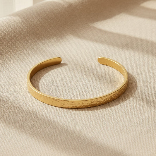 Goldfarbener Martella Armreif mit gehämmerter Oberfläche auf warmem beigem Leinenhintergrund.