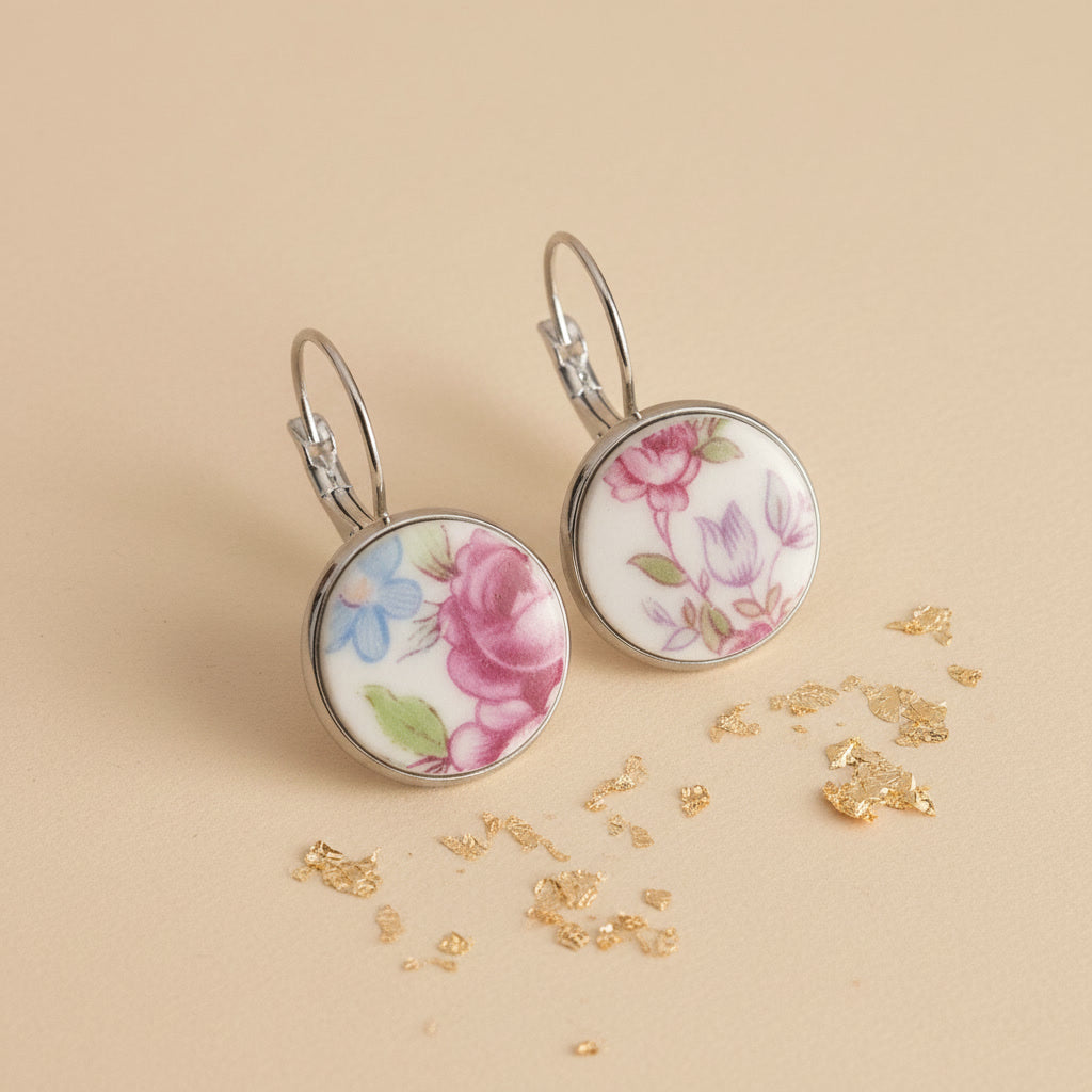 Seitenansicht der Floris Porzellan-Ohrringe von Edelatelier, die die hochwertigen Klappbrisuren aus Edelstahl zeigen. Die runden Inlays aus Upcycling-Porzellan präsentieren eine abwechslungsreiche Blütenpracht mit einer großen pinkfarbenen Rose links und filigranen violetten sowie hellblauen Blüten rechts. Das Paar ist auf einem beigefarbenen Hintergrund mit dekorativen Goldflocken und sanfter Schattenbildung arrangiert.