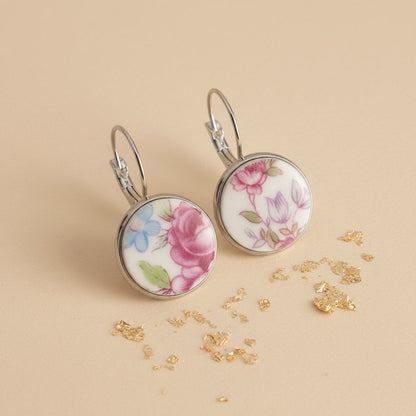 Seitenansicht der Floris Porzellan-Ohrringe von Edelatelier, die die hochwertigen Klappbrisuren aus Edelstahl zeigen. Die runden Inlays aus Upcycling-Porzellan präsentieren eine abwechslungsreiche Blütenpracht mit einer großen pinkfarbenen Rose links und filigranen violetten sowie hellblauen Blüten rechts. Das Paar ist auf einem beigefarbenen Hintergrund mit dekorativen Goldflocken und sanfter Schattenbildung arrangiert.