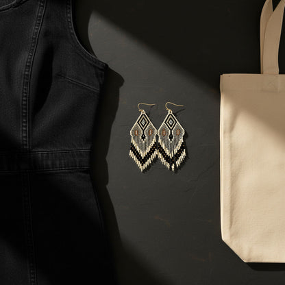 Eine edle Nahaufnahme im Flatlay-Stil zeigt die handgefertigten Perlenohrringe von EDELATELIER. auf einer dunklen Schieferplatte. Das elegante Design in Creme, Grau und Schwarz wird durch goldfarbene Akzente ergänzt. Links im Bild ist ein schwarzes Kleidungsstück aus Denim zu sehen, während rechts eine beige strukturierte Handtasche ins Bild ragt. Ein gezielter Lichteinfall hebt die feine Textur der gewebten Perlen und die hochwertige Verarbeitung der Statement-Ohrringe hervor.