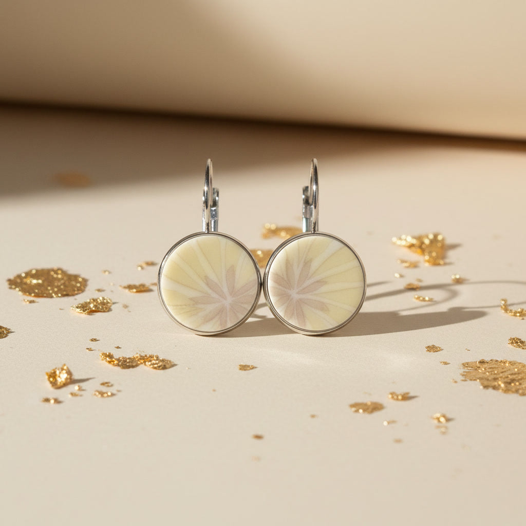 Nahaufnahme der kreisrunden Porzellanohrringe Modell Radiata von EDELATELIER.. Das Motiv zeigt ein strahlend gelbes, florales Muster, das sich wie Sonnenstrahlen über das reinweiße, glänzende Porzellan ausbreitet. Die Ohrringe sind in einer schmalen, silberfarbenen Edelstahlfassung sicher gehalten und liegen auf einer sandfarbenen Oberfläche im warmen Atelierlicht.