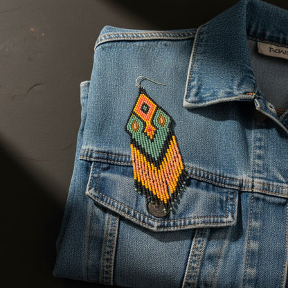 Die grün-ockergelben Perlenohrringe liegen dekorativ auf einer hellblauen Denim-Jacke. Der Kontrast zwischen dem blauen Stoff und den erdigen Naturtönen des Schmucks betont den modernen Boho-Look der Unikate.
