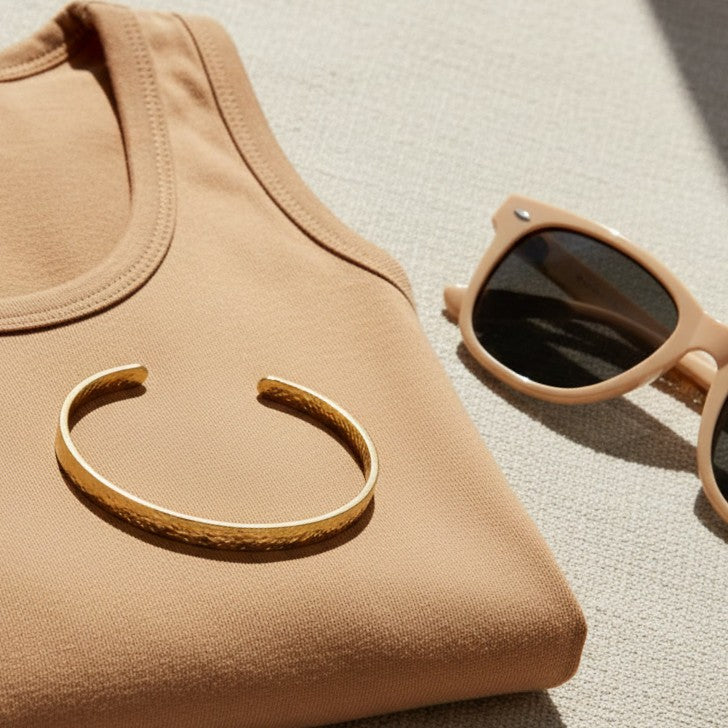 Goldfarbener Martella Armreif auf beigem Top neben Sonnenbrille.