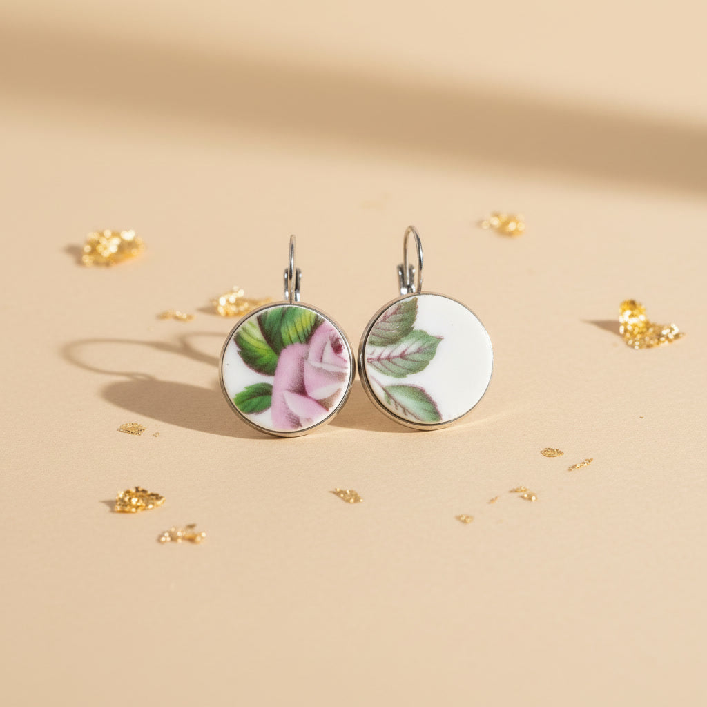 Handgefertigte Porzellanohrringe Memoria mit zartem rosa Blumendekor, liegend auf beigem Hintergrund mit Goldflocken von EDELATELIER.