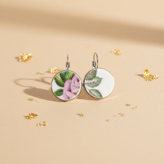 Handgefertigte Porzellanohrringe Memoria mit zartem rosa Blumendekor, liegend auf beigem Hintergrund mit Goldflocken von EDELATELIER.