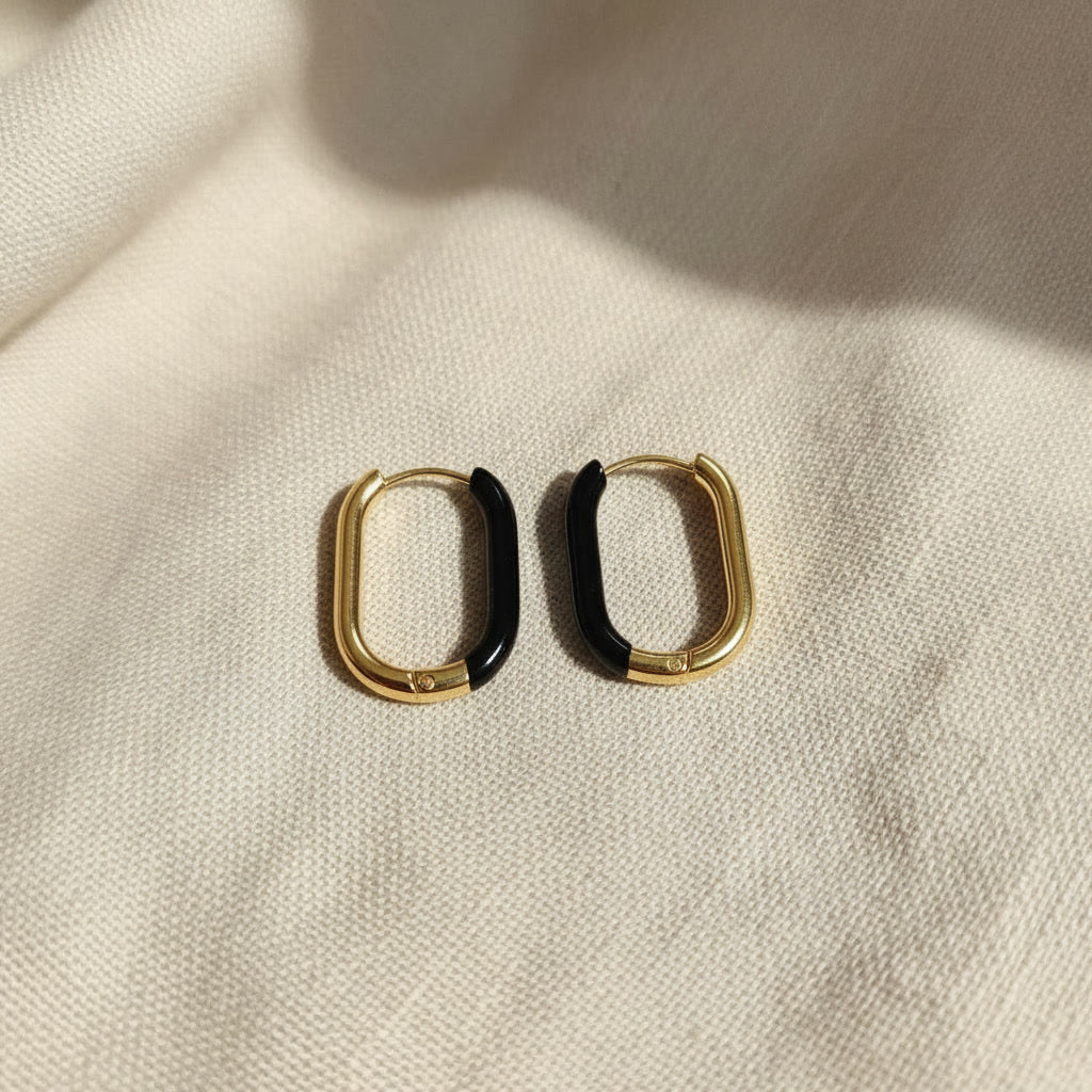 Nera Creolen in Gold und Schwarz frontal auf beigem Leinen, ovale Form und schwarzer Resin-Akzent sichtbar.