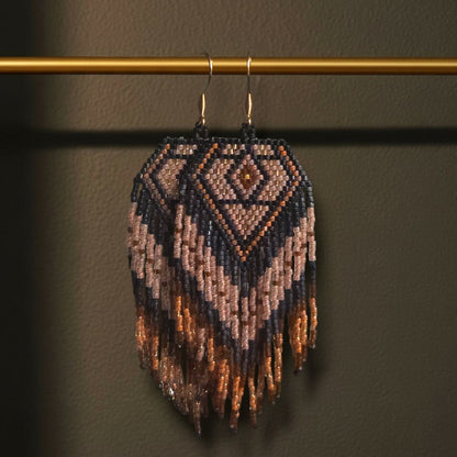 Handgewebte Perlenohrringe Notte Dorata an einer goldenen Stange vor einem dunklen Hintergrund. Das Design zeigt ein tiefblaues Rautenmuster mit edlen Goldakzenten und langen Fransen.