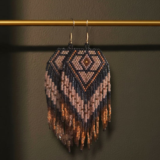 Handgewebte Perlenohrringe Notte Dorata an einer goldenen Stange vor einem dunklen Hintergrund. Das Design zeigt ein tiefblaues Rautenmuster mit edlen Goldakzenten und langen Fransen.