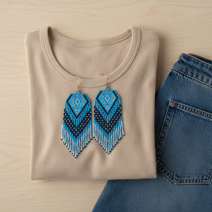 Ein helles Flatlay-Foto zeigt die blauen Perlenohrringe von EDELATELIER. platziert auf einem beigefarbenen T-Shirt neben einer blauen Jeans. Die Farbkombination unterstreicht den maritimen und sommerlichen Charakter des Designs.