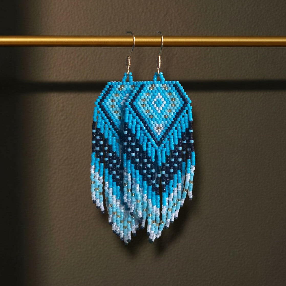 Eine Nahaufnahme der handgefertigten Perlenohrringe „Oceano“ von EDELATELIER., die elegant an einer goldenen Stange hängen. Das Design besticht durch ein geometrisches Rautenmuster in kräftigem Türkis, tiefem Nachtblau und reinem Weiß mit fließenden Perlenfransen am unteren Ende.