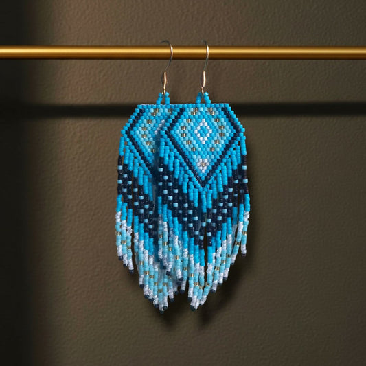 Eine Nahaufnahme der handgefertigten Perlenohrringe „Oceano“ von EDELATELIER., die elegant an einer goldenen Stange hängen. Das Design besticht durch ein geometrisches Rautenmuster in kräftigem Türkis, tiefem Nachtblau und reinem Weiß mit fließenden Perlenfransen am unteren Ende.
