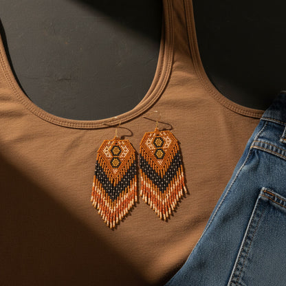 Ein stimmungsvolles Flatlay von EDELATELIER. zeigt die handgefertigten Perlenohrringe „Oro Bruno“ auf einem braunen Tanktop, flankiert von einer blauen Jeans. Das Design der Ohrringe besticht durch warme Erdtöne wie Ocker, Dunkelbraun und Beige, die in einem komplexen geometrischen Muster mit fließenden Fransen gewebt sind. Ein markantes Spiel aus Licht und tiefen Schatten verleiht der Szene eine edle, warme Atmosphäre und betont die feine Textur der Miyuki-Perlen.